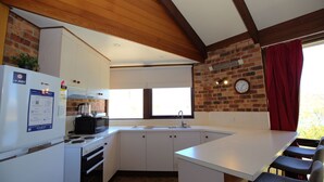 Una cocina compacta con armarios blancos, un microondas integrado, una estufa y un lavabo. Hay una ventana con cortina roja, un reloj en la pared y una planta en la encimera.