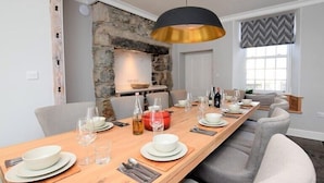 A dining area with a long wooden table set for six, a stone wall, a modern pendant light, and a view of the outside through a window.