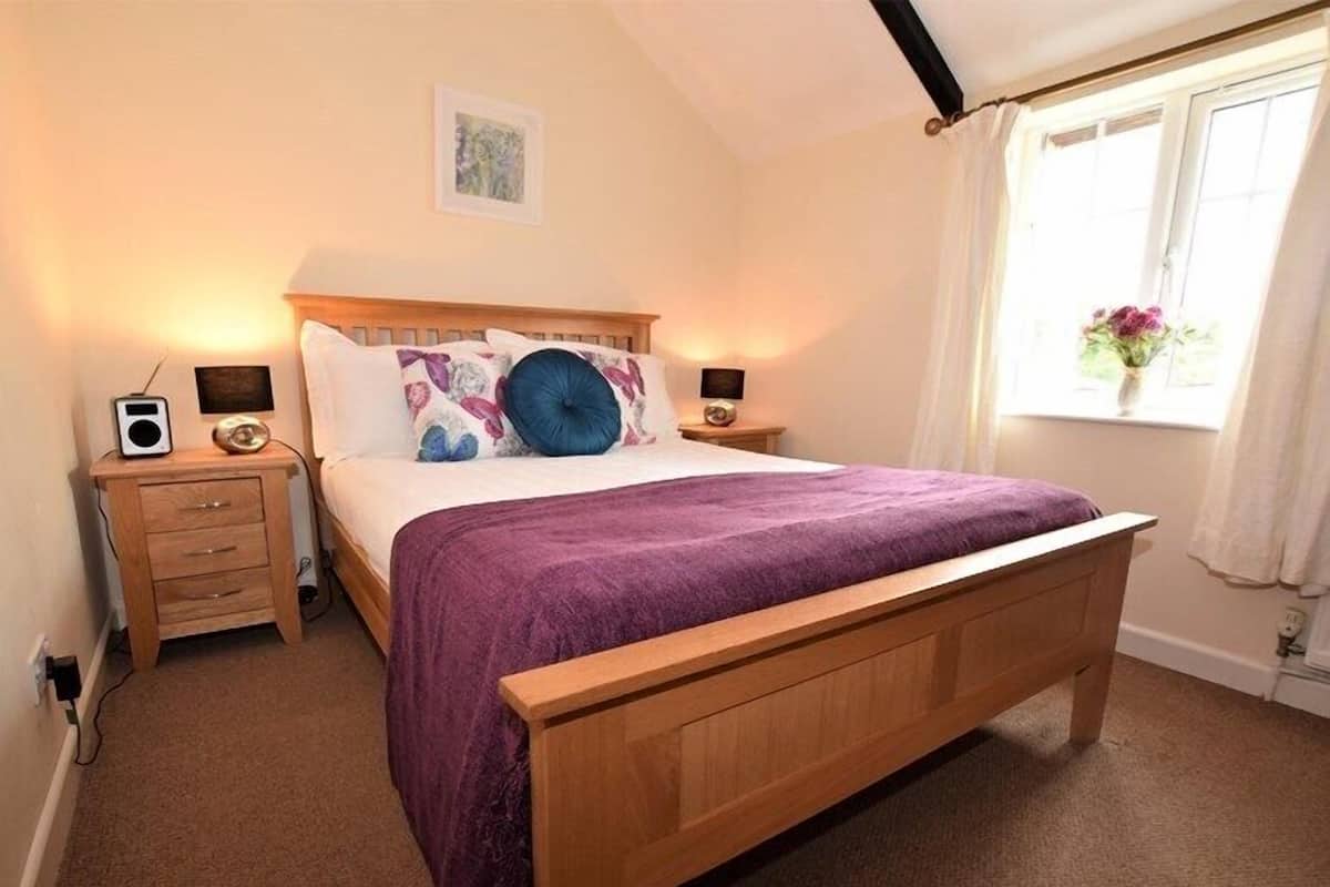 A bedroom with a wooden bed, purple bedding, two bedside tables with lamps, a small radio, and a window with curtains.