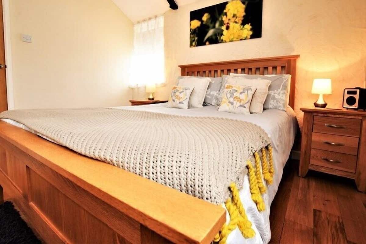 A bedroom with a wooden bed, a bench at the foot of the bed, a nightstand with a lamp, and a painting on the wall.