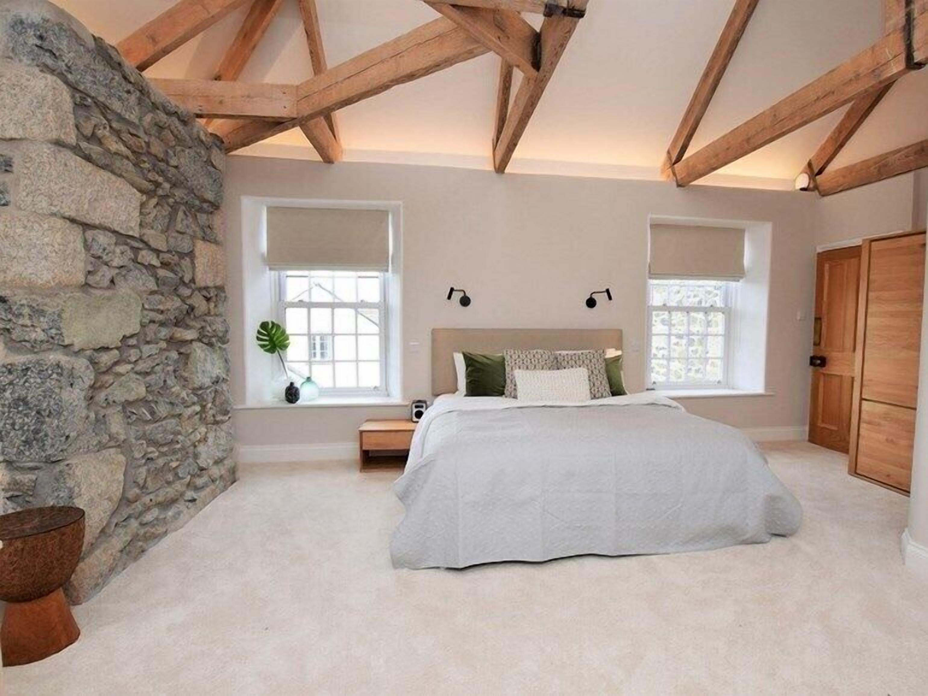 Une chambre avec un mur en pierre, un lit avec une literie blanche, deux fenêtres équipées de stores et des poutres en bois au plafond.