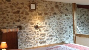 A room with a stone wall, a bed with a patterned bedspread, a wooden headboard, a nightstand with a lamp, and a window with a wooden frame. - Beautiful renovated stone barn with magnificent view! Quiet village (Rabouillet)