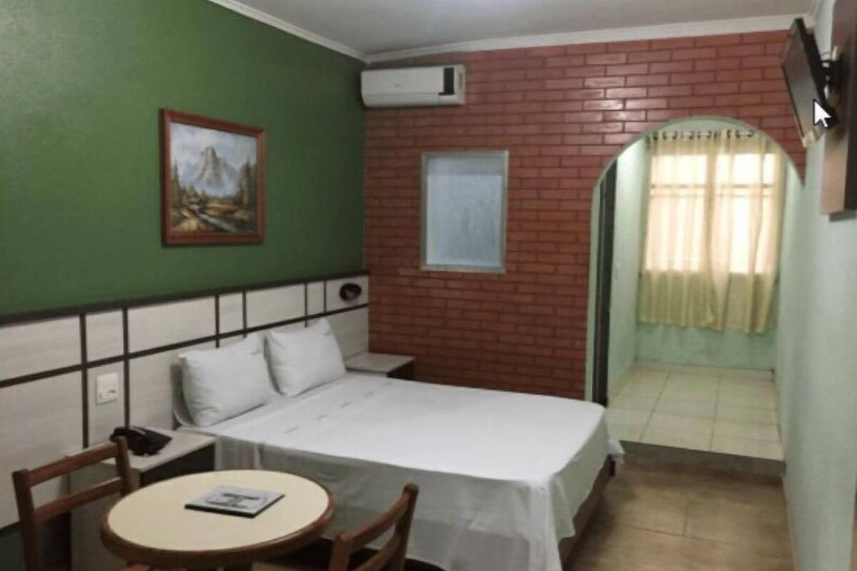 Une petite chambre avec un lit simple, un mur vert, une fenêtre avec des rideaux, une petite table avec une chaise et un tableau accroché au mur.