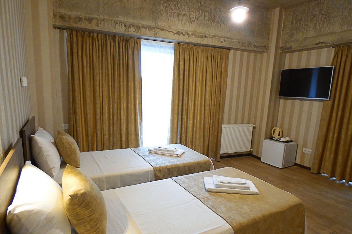 A hotel room with two single beds, a flat-screen TV, a small bedside table with a lamp, and a window with curtains.