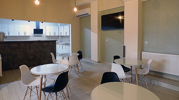 A modern dining area with round tables and chairs, a kitchen in the background, and a large window.