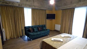 A room with a green sofa, two beds with white linens, wooden flooring, a flat-screen TV on the wall, and large windows with curtains.