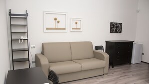 Una habitación con un sofá color beige, un escritorio negro con una silla, un aparador negro, un radiador blanco y dos cuadros en la pared.