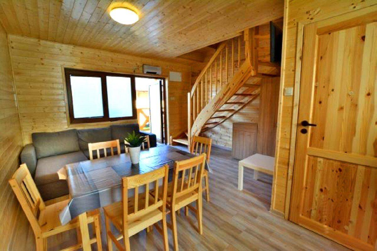 A wooden cabin interior with a dining table, chairs, a sofa, a staircase, and a large window.