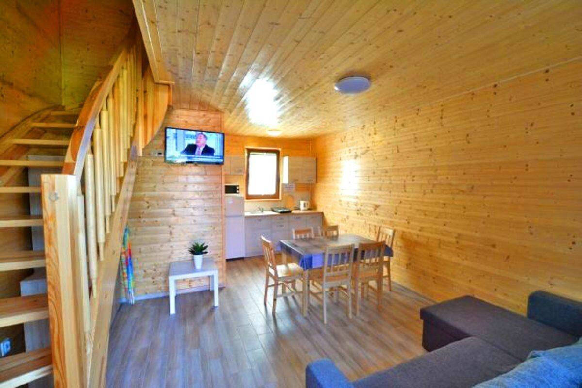 A wooden cabin interior with a dining area, a sofa, a TV mounted on the wall, and a staircase.
