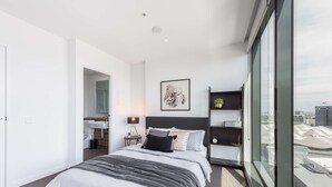 A modern bedroom with a large bed, a bedside table with a lamp, a framed artwork on the wall, and a view of the city through large windows. - Open View towards Stadium - Southern Cross 2B2Bath (Melbourne)