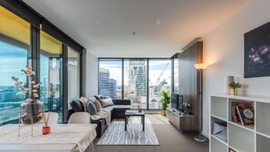 A modern living room with a sofa, coffee table, and TV, large windows with a city view, and a bookshelf. - Open View towards Stadium - Southern Cross 2B2Bath (Melbourne)