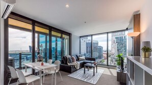 A modern living room with a sofa, dining table, and chairs. It features large windows with a city view. - Open View towards Stadium - Southern Cross 2B2Bath (Melbourne)