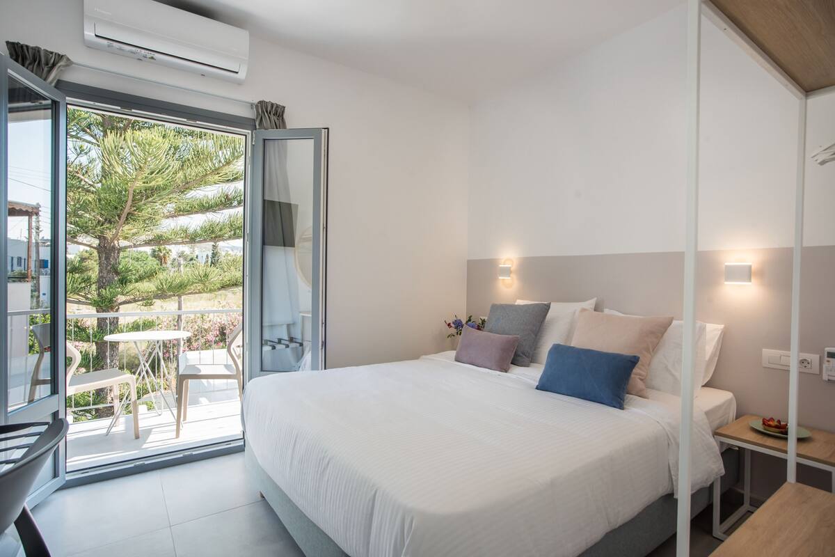 A modern bedroom with a large bed, a balcony with a table and chairs, and a view of greenery.