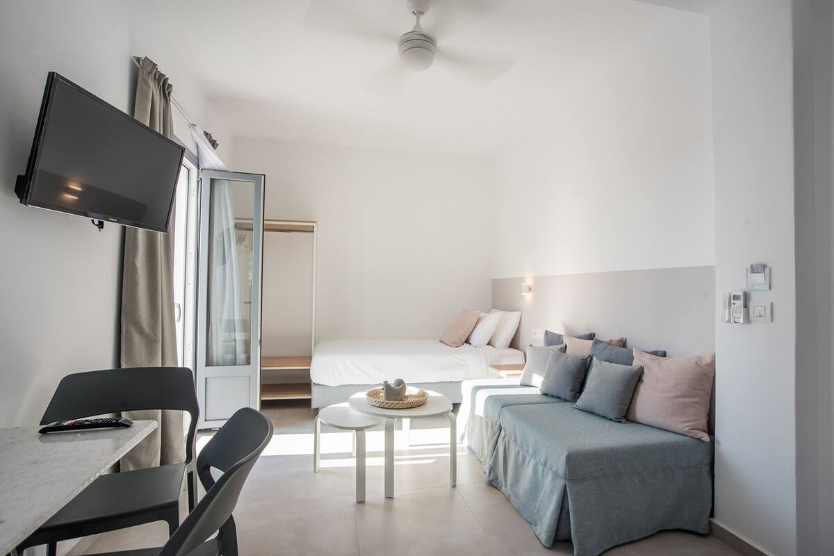 A modern hotel room with a bed, a sofa, a TV, a table, and a ceiling fan.