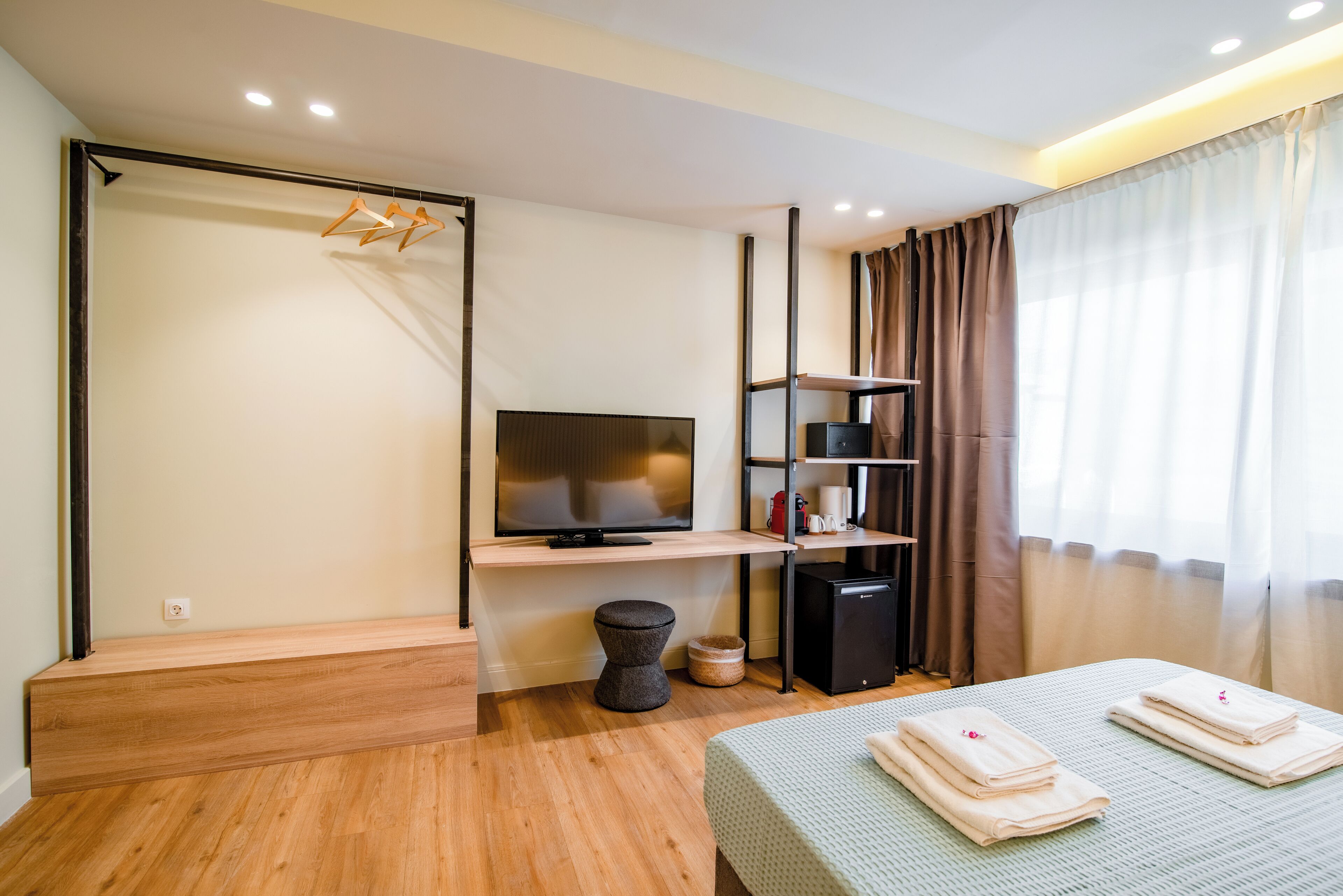 A modern hotel room with a wooden floor, a flat-screen TV, a small desk, a sofa with towels, a trash bin, and a wardrobe with hanging clothes.
