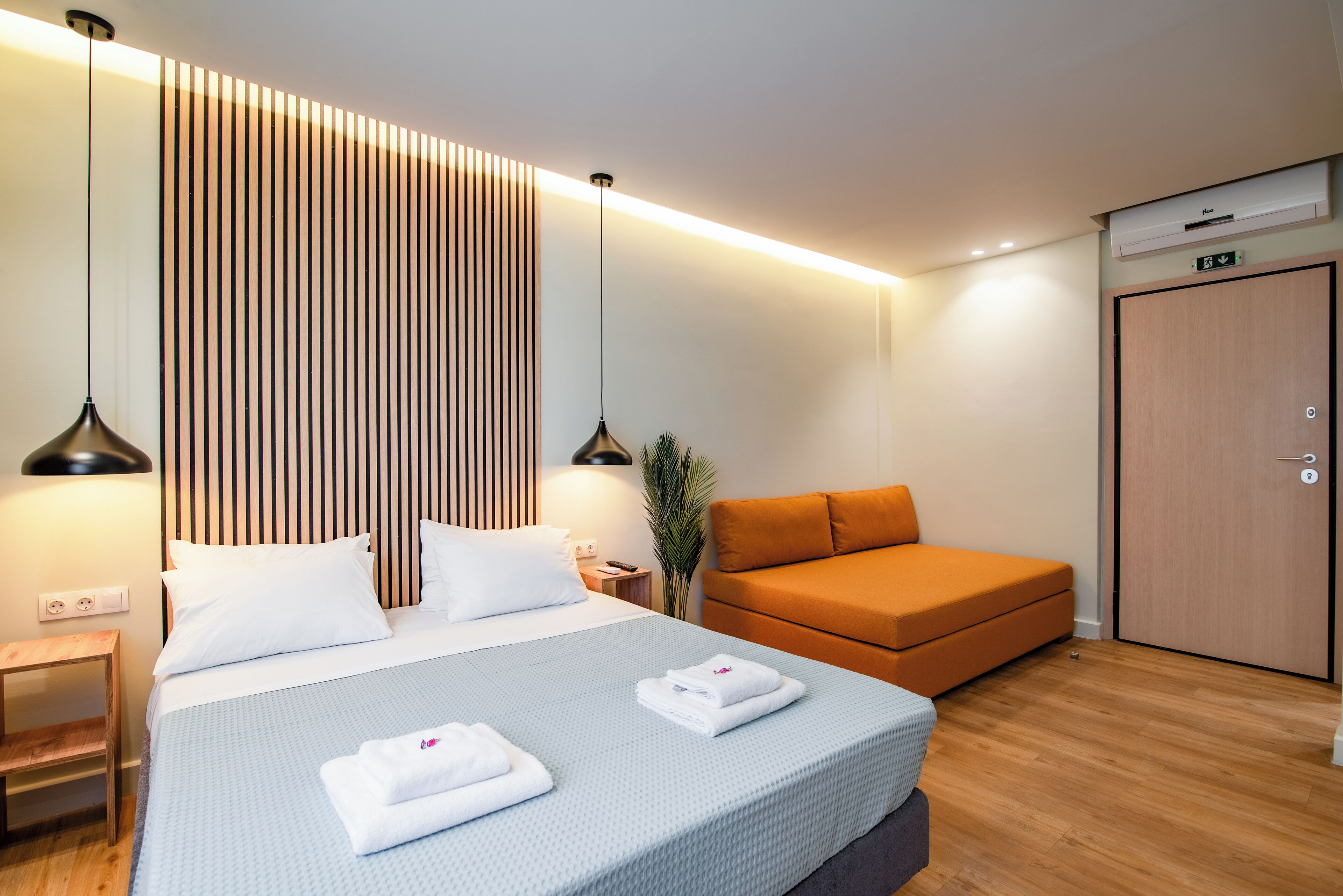 Una camera d'hotel moderna con un letto grande, un divano arancione, un pavimento in legno e un condizionatore d'aria a parete.