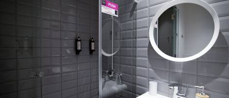 A modern bathroom with a push button for the shower, a round mirror, and a white sink with a wooden soap holder.