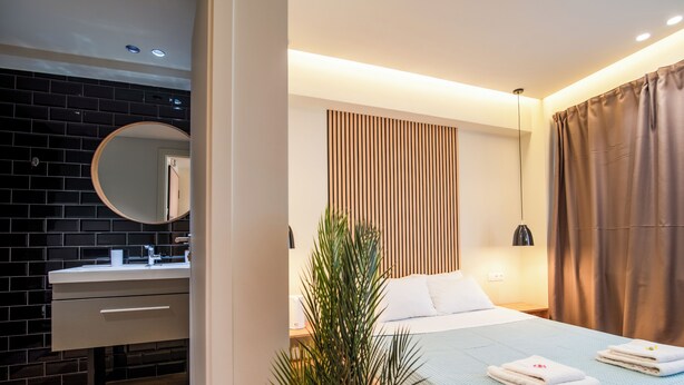 Una habitación de hotel moderna con una pared de ladrillo negro, un espejo redondo, una cama blanca con colcha azul y un panel de pared de listones de madera.