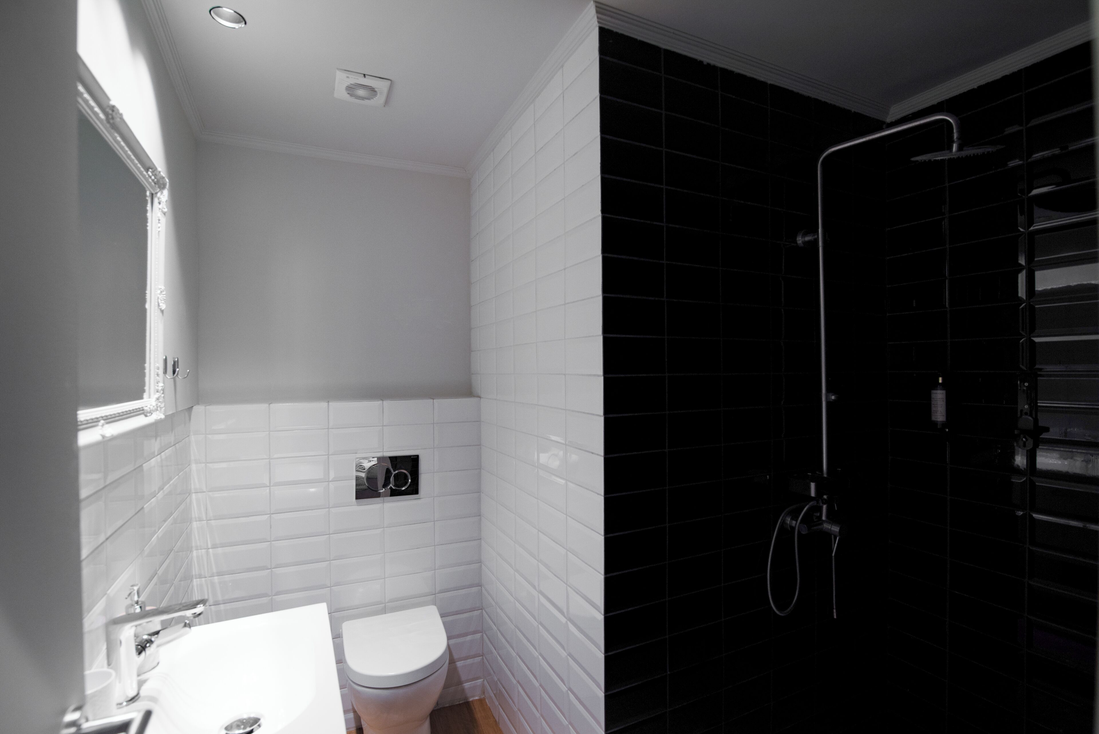 A bathroom with white tiled walls, a white sink, and a toilet. There is a shower area with black tiles and a glass door.