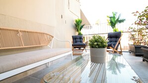 Una mesa con superficie de vidrio, un banco tejido y dos sillones, una planta en maceta y una barandilla de balcón.