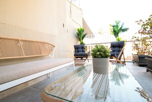 Un tavolo con piano in vetro, una panca intrecciata e due poltrone, una pianta in vaso e la ringhiera di un balcone.