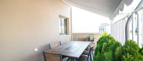 An outdoor dining area with a long table and chairs, a small window, and a view of buildings in the background.