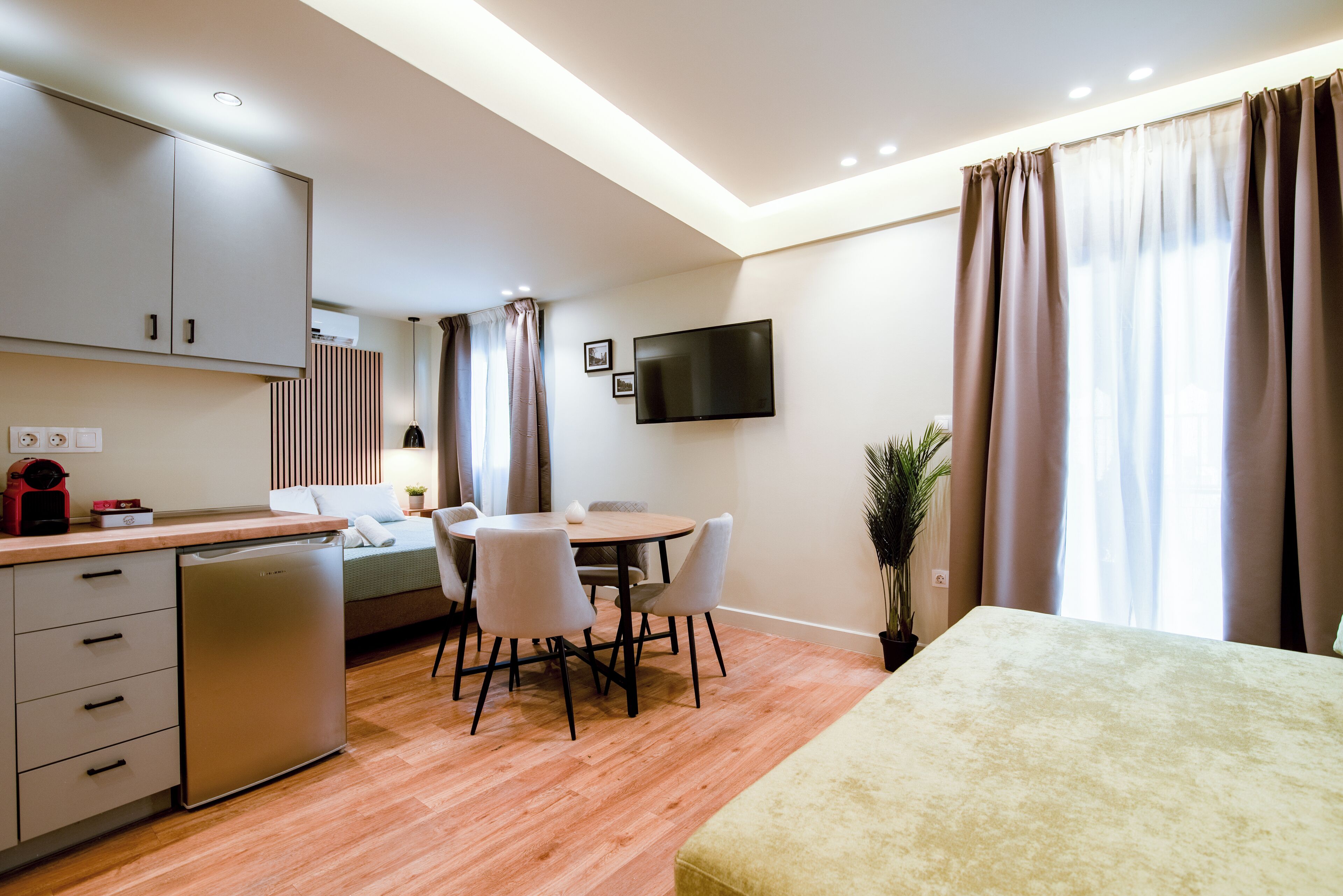 A modern kitchen with a dishwasher, a dining area with a round table and chairs, a bed with a headboard, and a TV mounted on the wall.