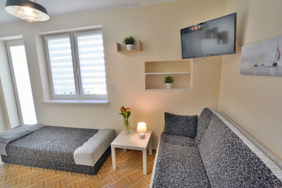 Ein modernes Wohnzimmer mit einer Couch, einem kleinen Tisch mit Lampe, einem an der Wand montierten Fernseher und einem Fenster mit Jalousien.