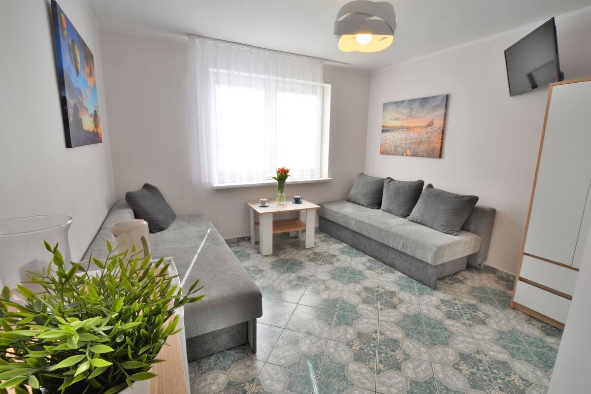 Ein modernes Wohnzimmer mit einem grauen Sofa, einem kleinen weißen Tisch mit einer Vase und einem großen Fenster mit transparenten Vorhängen.