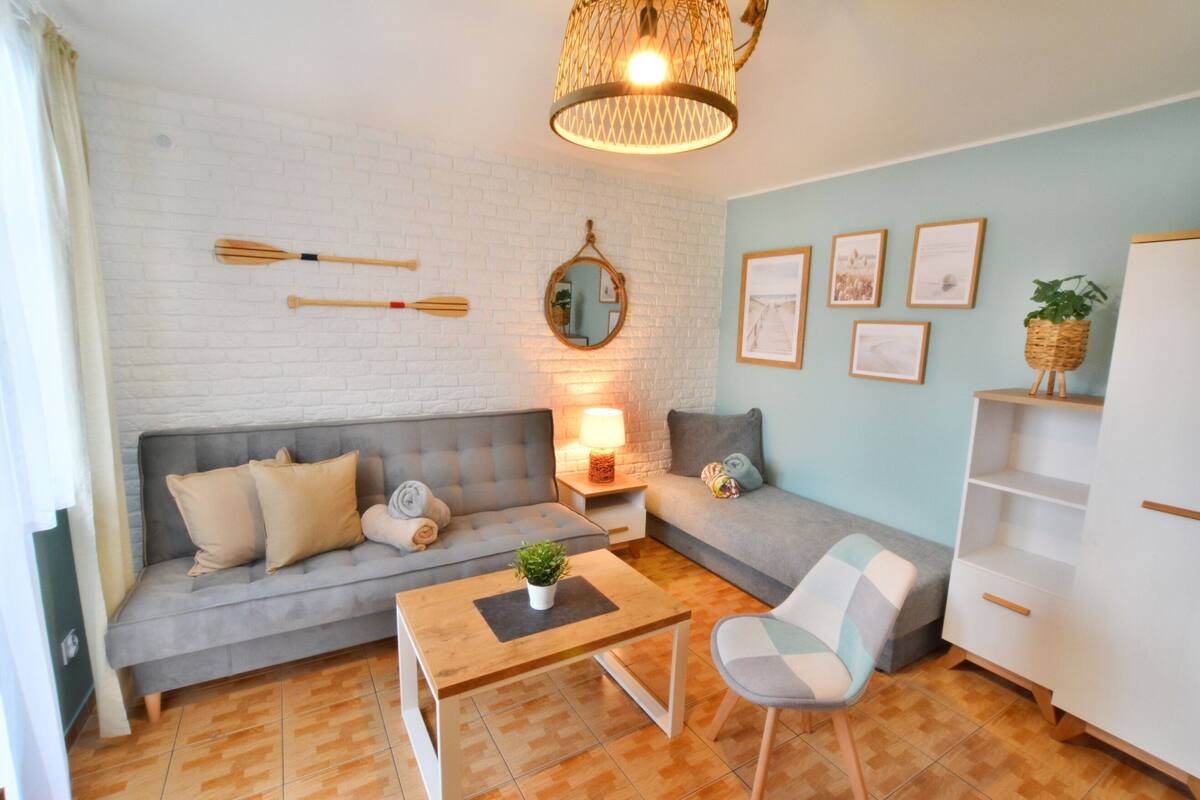 Ein Wohnzimmer mit grauem Sofa, einem Holztisch, einem Sessel, einem kleinen Tisch mit Lampe und einem wandmontierten Spiegel.