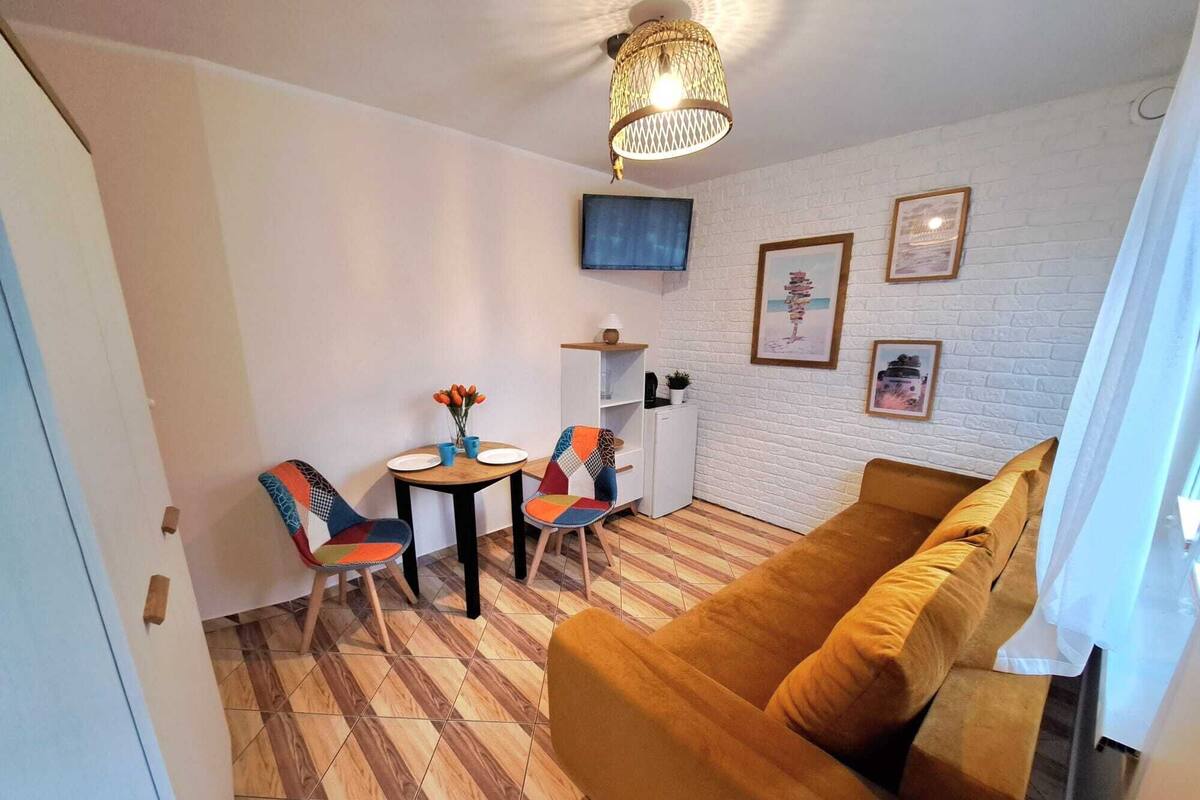 Ein Wohnzimmer mit einem braunen Sofa, zwei bunten Sesseln, einem kleinen Tisch mit Blumen in einer Vase, einem weißen Kühlschrank und einem an der Wand montierten Fernseher.