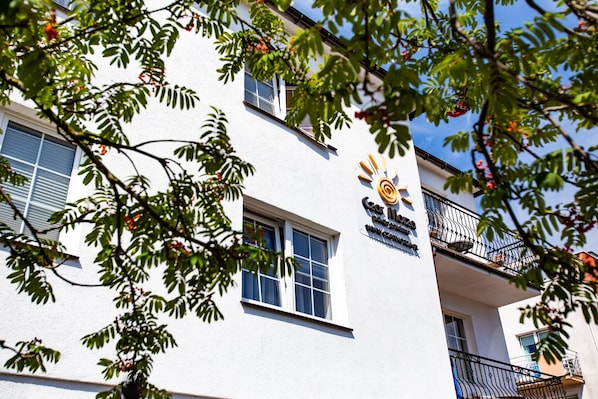 A building with a white facade, a balcony with black railings, and a sign with a sun and text.