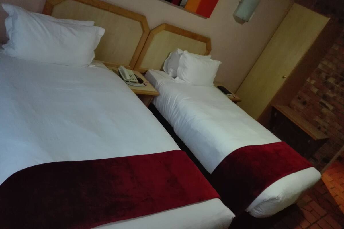A hotel room with two beds, each with white bedding and red throw pillows. There are bedside tables with phones and lamps.