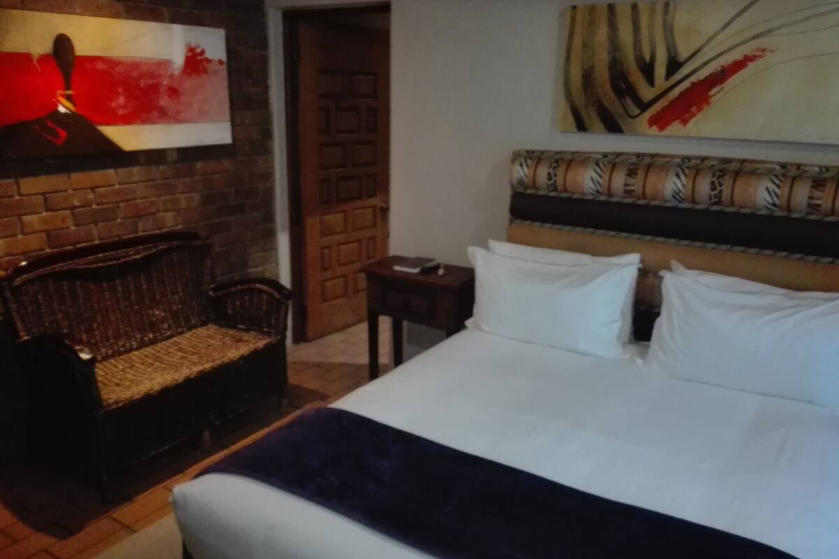 A hotel room with a large bed, a wooden headboard, a wicker chair, a small table, and a painting on the wall.