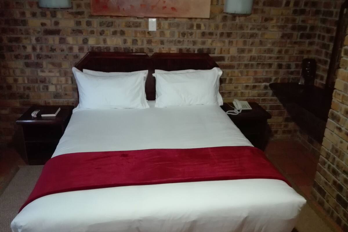A hotel room with a brick wall, a bed with white bedding and a red blanket, two bedside tables with lamps, and a phone on one table.