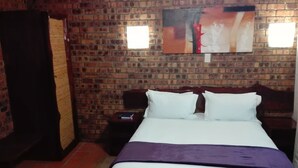 Une chambre d’hôtel avec un mur en briques, un lit avec une literie blanche et violette, une table de chevet et une petite table.