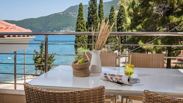A balcony with wicker furniture, a white pitcher, and a glass of drink, overlooking a coastal view with trees and mountains. - Meliti Waterfront Suites (Kefalonia)