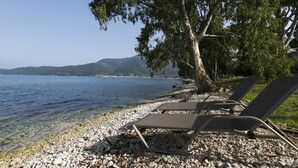 A pebble beach with lounge chairs and a tree, overlooking a calm lake and distant hills. - Meliti Waterfront Suites (Kefalonia)