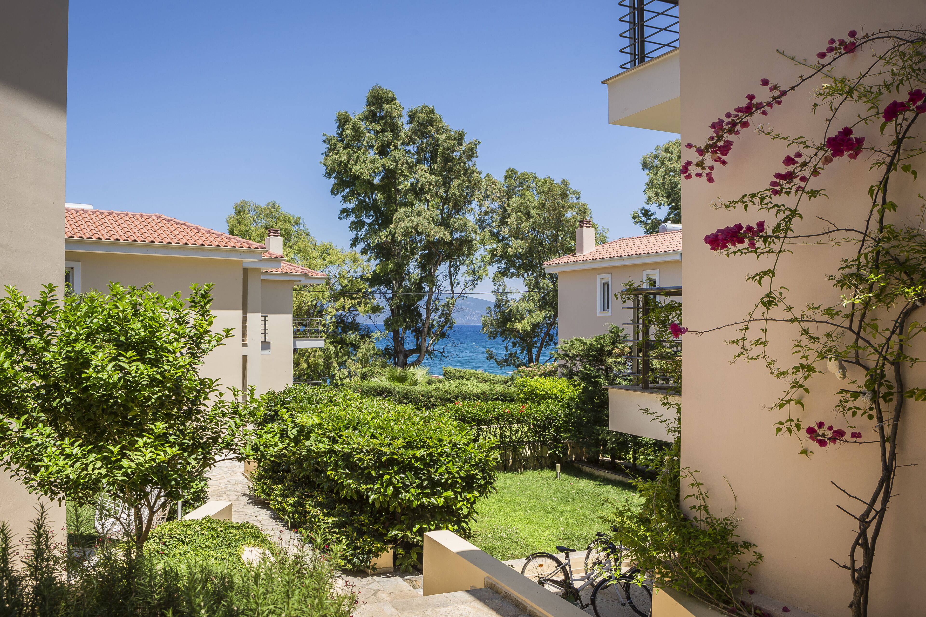 A courtyard with a view of the sea, surrounded by greenery and buildings with red-tiled roofs.