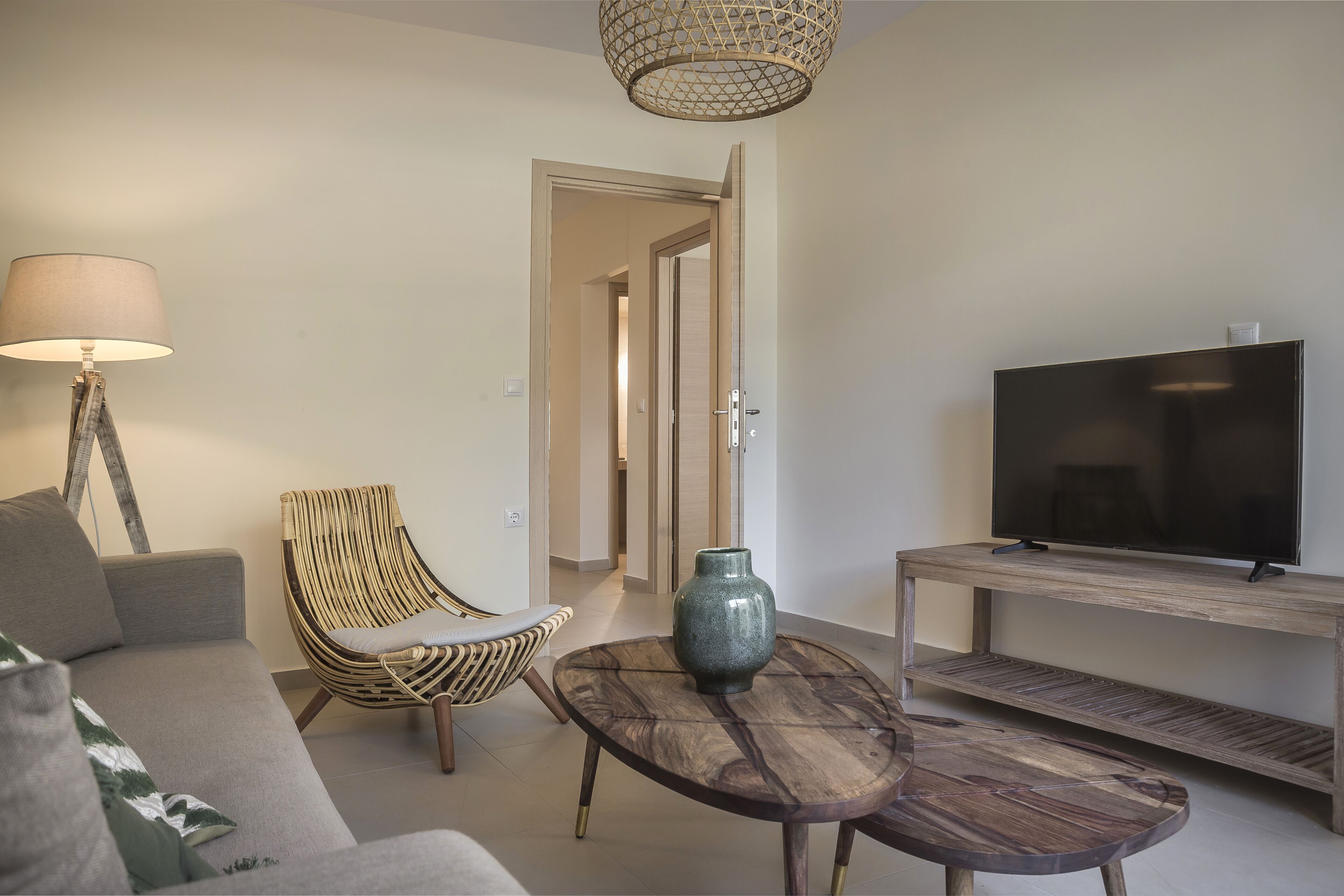 Una sala de estar con un sofá, una silla de mimbre, una mesa redonda de centro con un jarrón, una lámpara y un televisor sobre un soporte de madera.