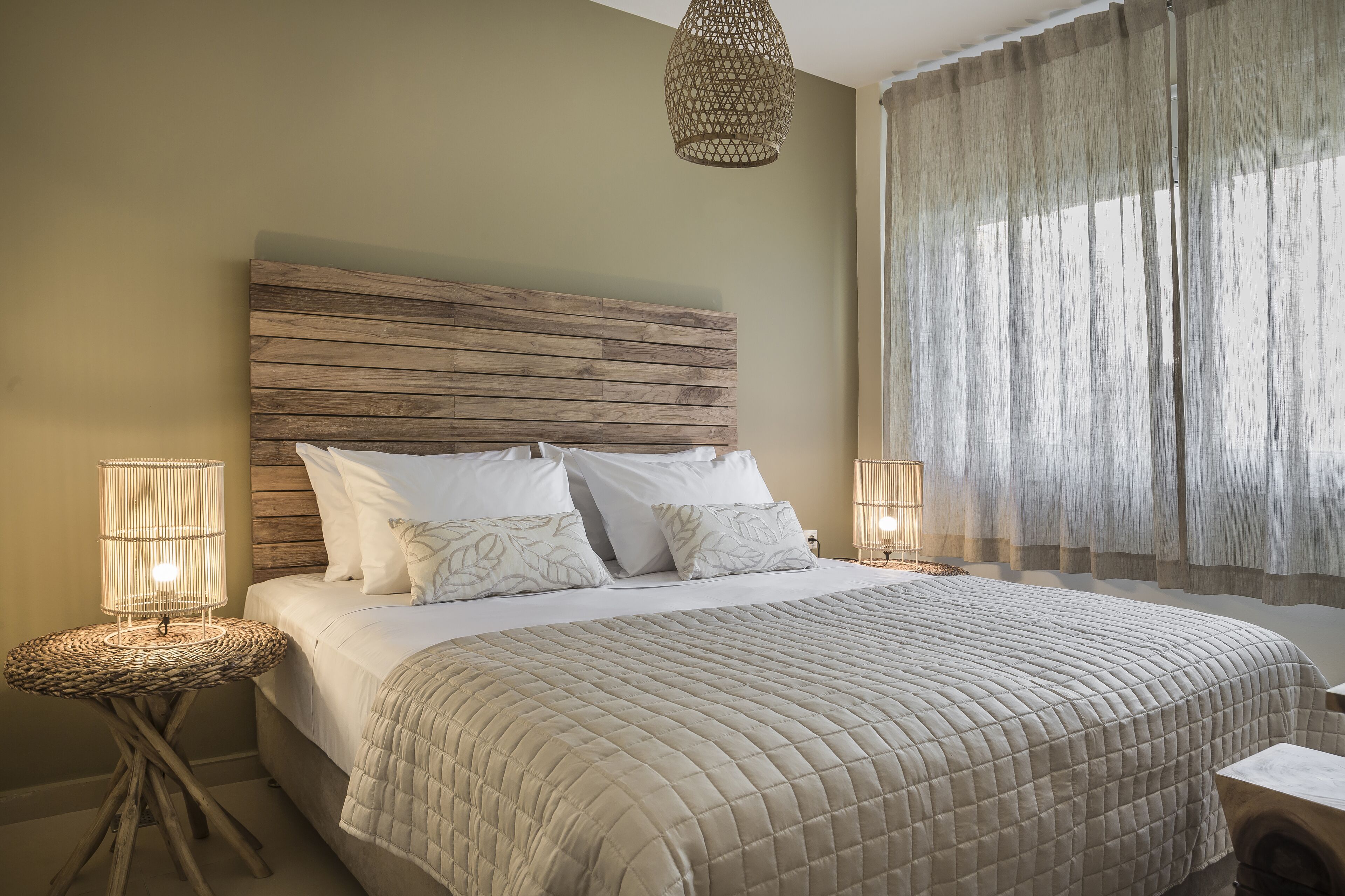 Quarto com cabeceira de madeira, cama com roupa de cama branca, duas luminárias de cabeceira e uma janela grande com cortinas leves.