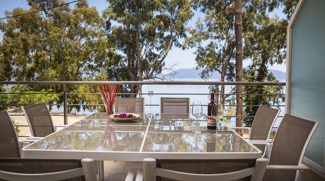 A glass-top table with chairs, a bottle, and a bowl of fruit on a balcony overlooking a body of water and trees.