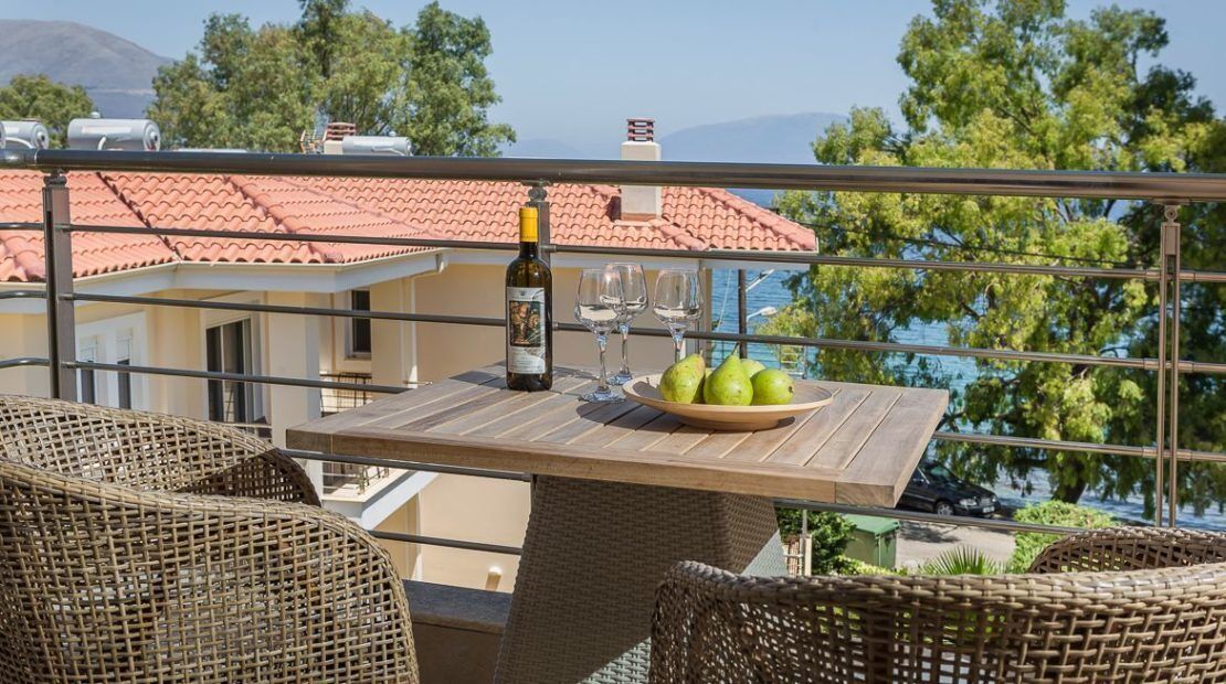 Ein Balkon mit Korbmöbeln, einem Tisch mit einer Weinflasche, Gläsern und Birnen, mit Blick auf ein rotes Ziegeldach und Bäume.