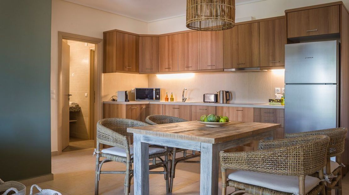 A modern kitchen with wooden cabinets, a stainless steel refrigerator, a wooden dining table with wicker chairs, and a hanging light fixture.