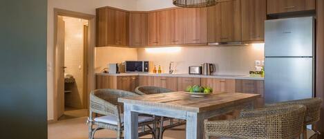 A modern kitchen with wooden cabinets, a stainless steel refrigerator, a wooden dining table with wicker chairs, and a hanging light fixture.