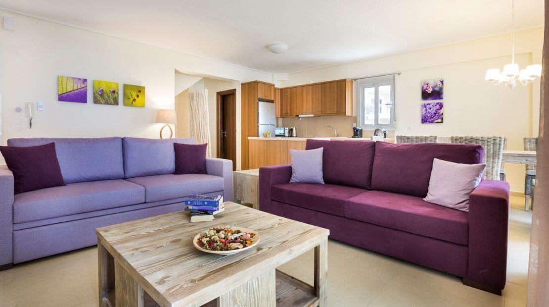 Un salon avec deux canapés violets, une table basse en bois et une cuisine en arrière-plan.