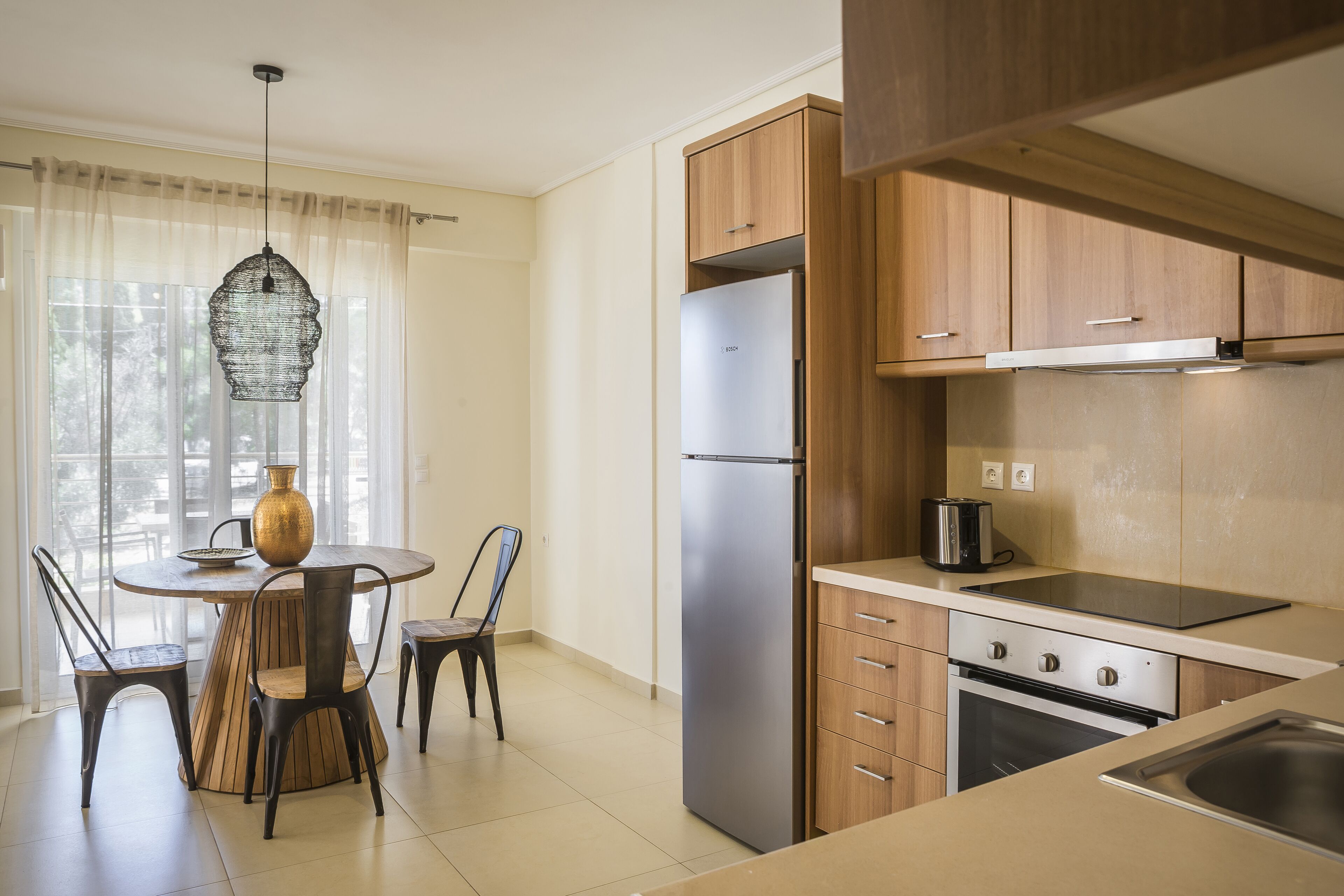 A modern kitchen with a stainless steel refrigerator, wooden cabinets, a dining table with chairs, and a hanging light fixture.