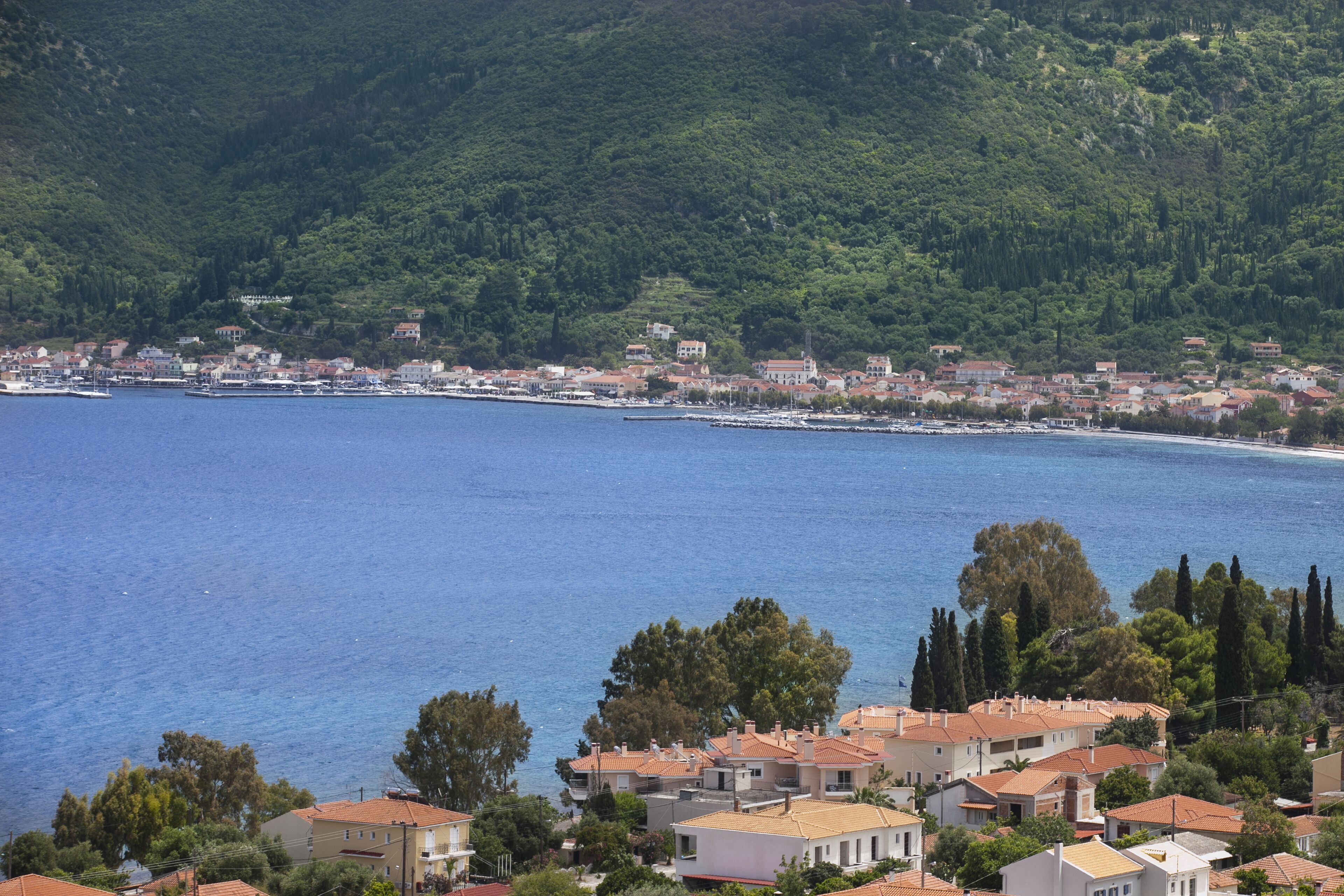A coastal town with buildings along the waterfront, surrounded by hills and dense greenery.