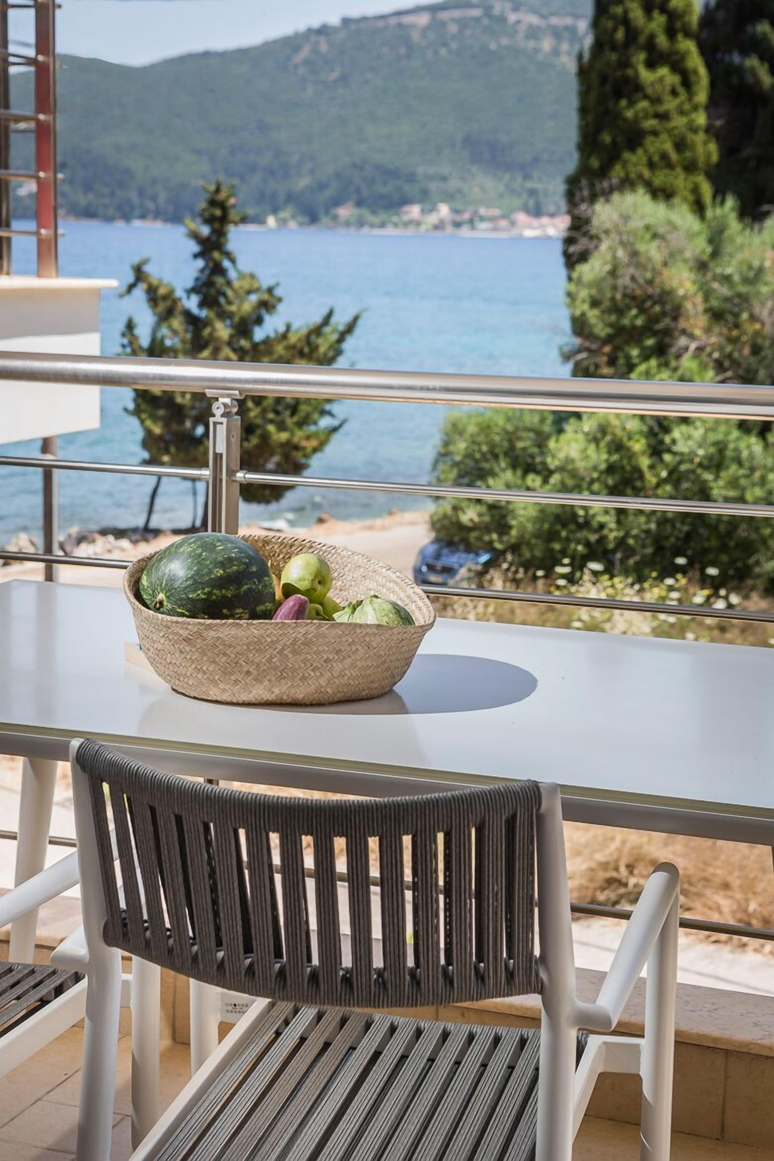 Une table sur laquelle repose un panier tressé contenant une pastèque et des pommes, une chaise en métal, et une vue sur un paysage côtier avec des arbres et une étendue d’eau.