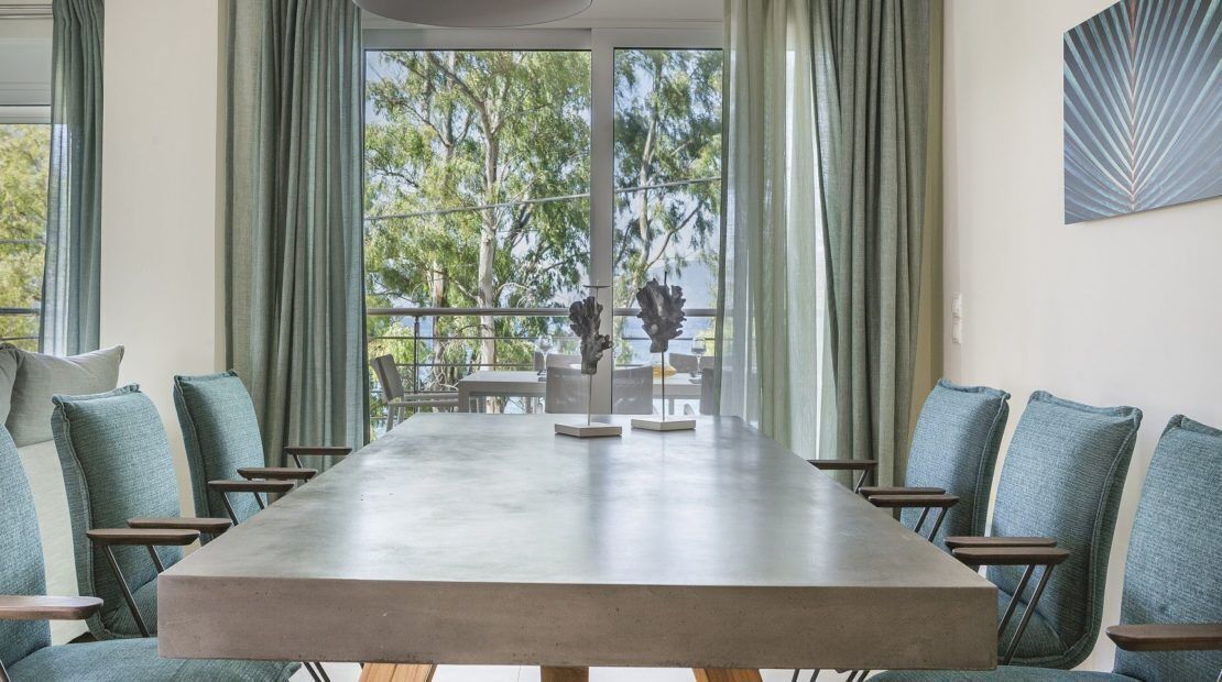 Un salón de conferencias con una larga mesa y sillas, ventanas amplias con cortinas y vistas a árboles.